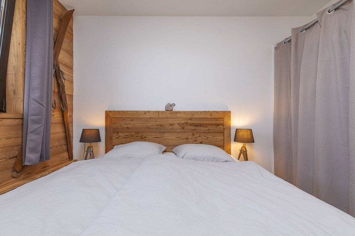 A cozy bedroom with a white bed, rustic wooden headboard, two lit lamps, wood-paneled wall, and vintage skis as decor.