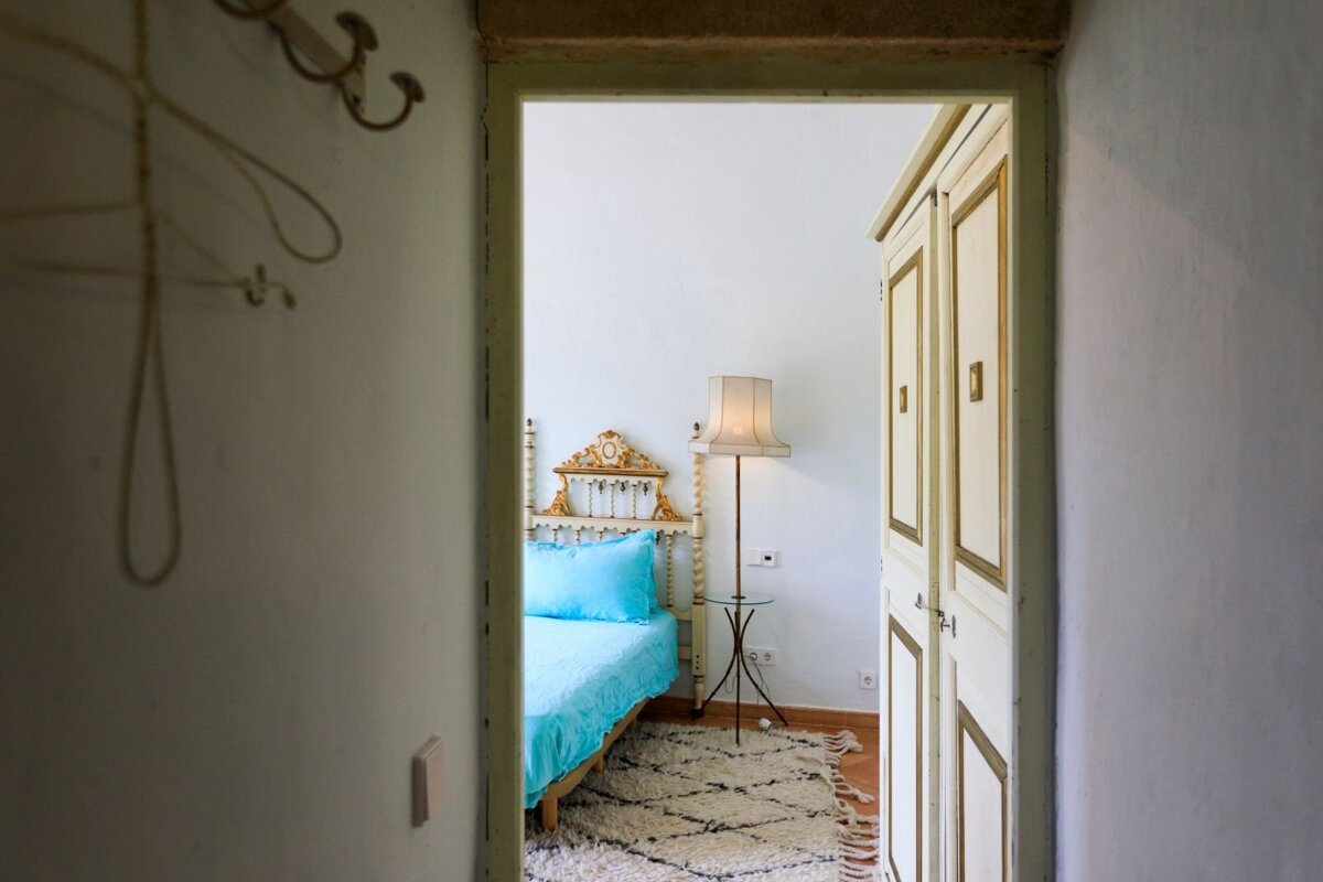 A bedroom seen through a doorway, featuring a blue bed with an ornate gold headboard, a floor lamp, white wardrobe, and a patterned rug.