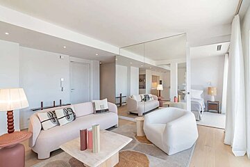 A bright, modern living room with a pink sofa, white armchair, and mirrored walls reflecting a kitchen area and a bedroom, creating a spacious feel.