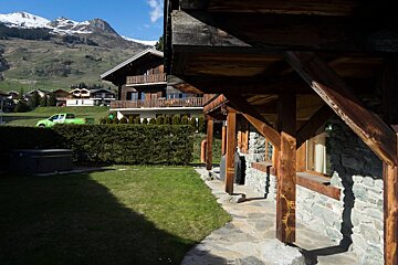 View from a rustic wooden chalet onto a green lawn, with traditional alpine houses and snowy mountains in the background.