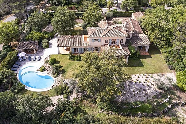 Aerial view of a sprawling villa with terracotta roofs, light blue shutters, a kidney-shaped pool, and lush green landscaping.