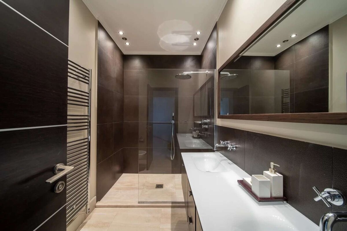 A modern bathroom with dark brown tiled walls, a white countertop, glass shower, and large mirror. Recessed lighting adds a warm, clean feel.