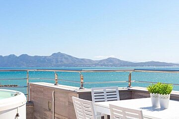 Sunny terrace with white outdoor furniture overlooking a bright blue sea and distant mountains under a clear sky.