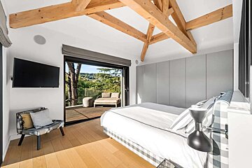 Modern bedroom with exposed timber beams, a large glass door opening to a balcony with a scenic green view, and light wood flooring.
