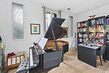 A well-lit room features a grand piano with sheet music, flanked by bookshelves, a cluttered desk, and decorative patterned windows.