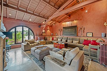Spacious living room with terracotta walls, exposed wooden beams, large sectional sofa, and an arched window overlooking a green landscape.