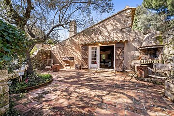 A sunny, rustic terracotta patio shaded by a large tree, adjacent to a stone house with open doors and an outdoor fireplace.