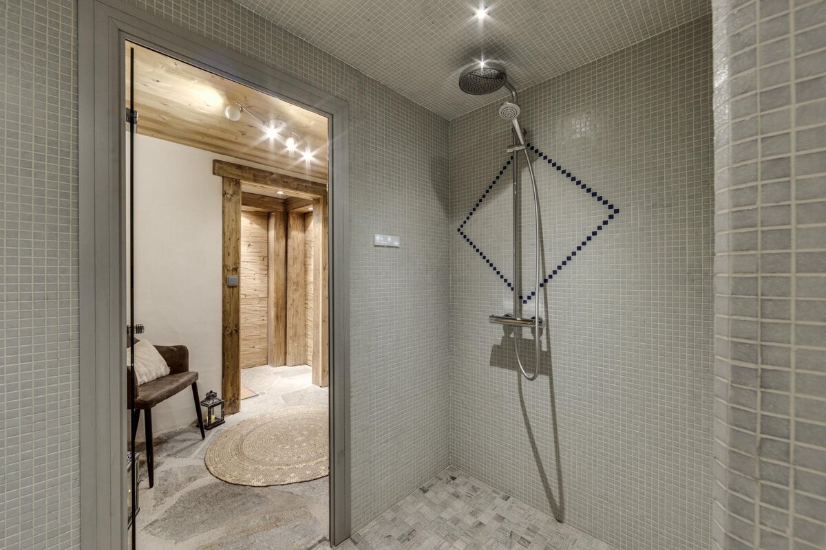 A modern shower stall with grey mosaic tiles and a blue diamond pattern, leading to a rustic room with wooden walls, stone floor, and a bench.