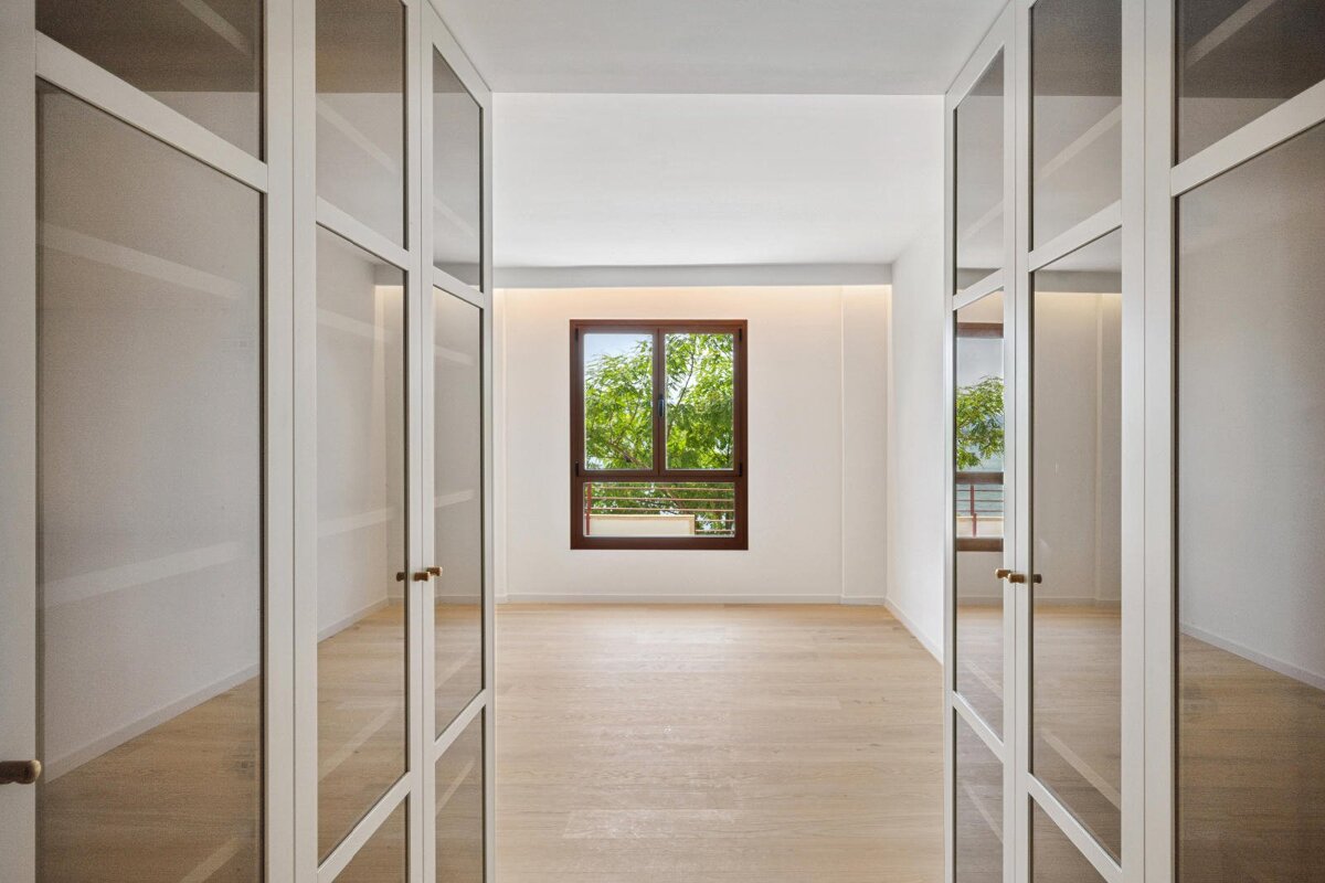 A minimalist room with light wood floors and white walls, viewed through glass doors. A large window at the back showcases lush green trees.