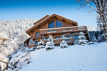 A magnificent wooden chalet blanketed in snow, nestled among snow-covered evergreen trees against a clear blue winter sky.