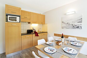 A compact, modern kitchen with light wood cabinets, integrated appliances, and a dining table set for four with ski-themed plates, wine, and bread.