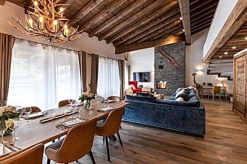 A cozy, luxurious chalet great room featuring a rustic wood-beamed ceiling, antler chandelier, stone fireplace, dining table, and blue sofa.