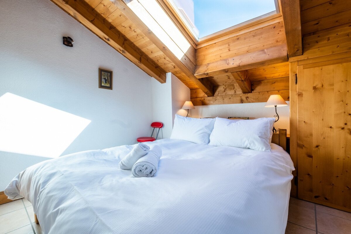 A bright, cozy attic bedroom features a white bed, wooden beamed ceiling, and a skylight. White walls, bedside lamps, and a red chair add charm.
