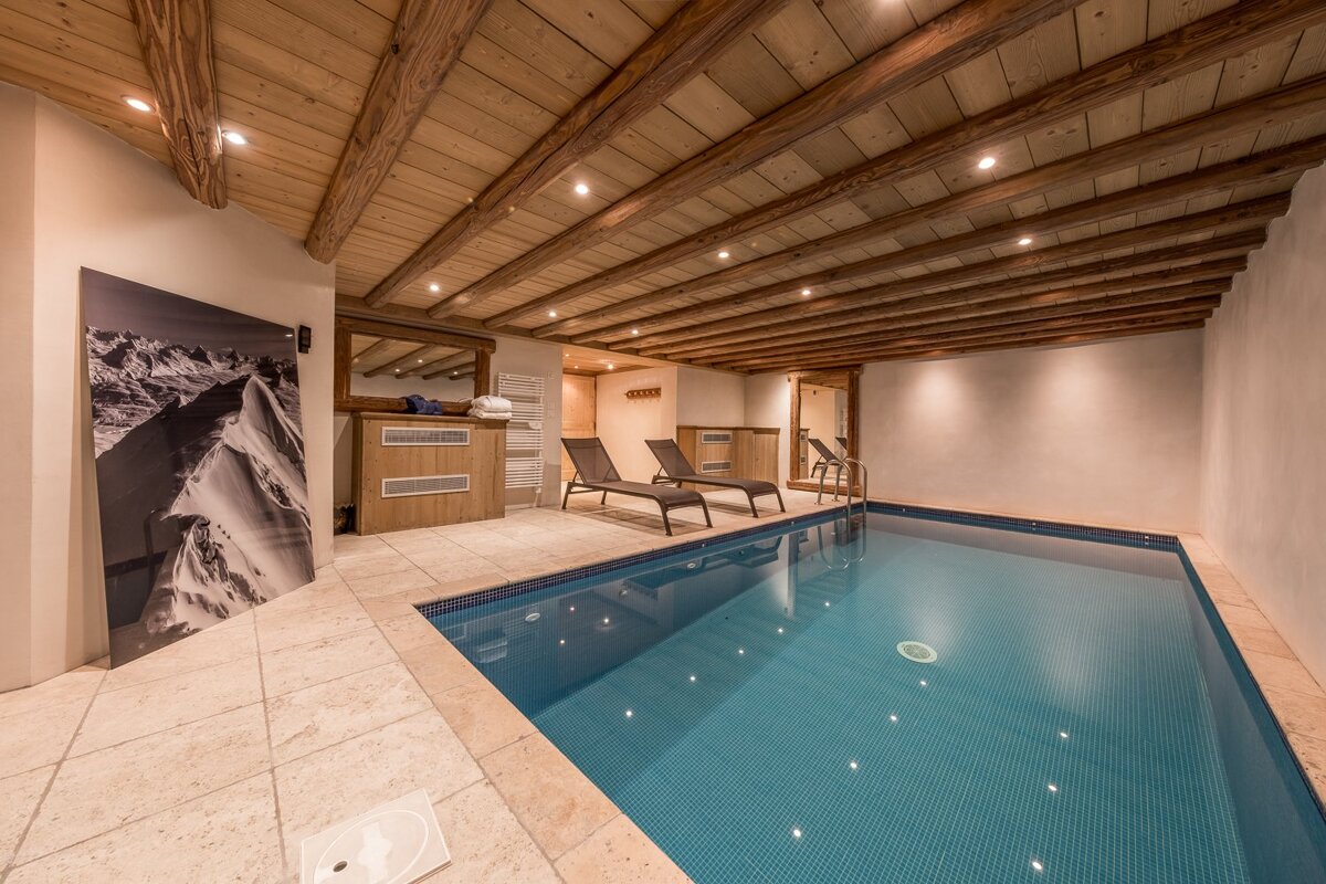 Cozy indoor pool with a rustic wood-beamed ceiling, tiled floor, lounge chairs, and mountain art, creating a relaxing spa-like ambiance.