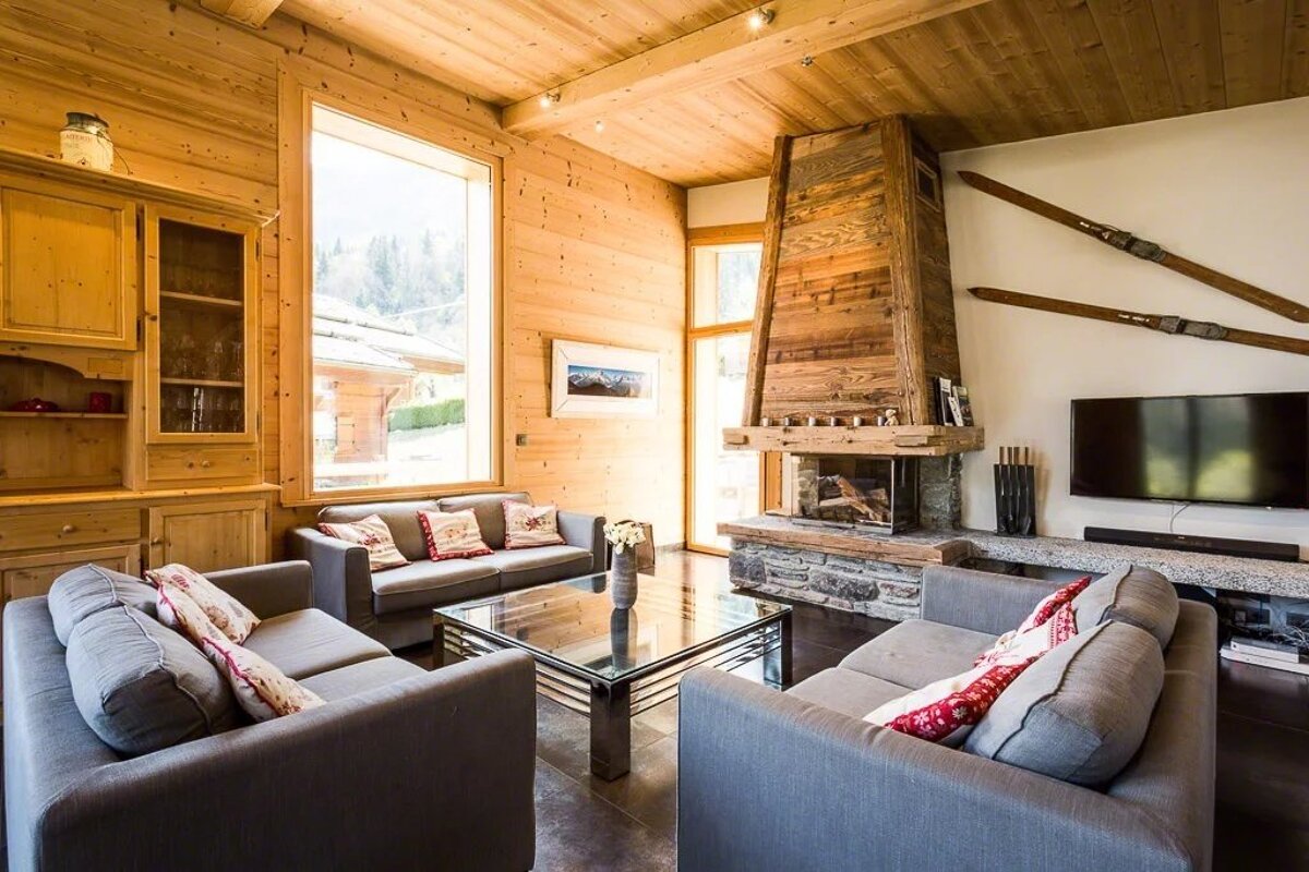 A cozy cabin living room with wooden walls, three grey sofas, a glass coffee table, and a rustic stone fireplace.