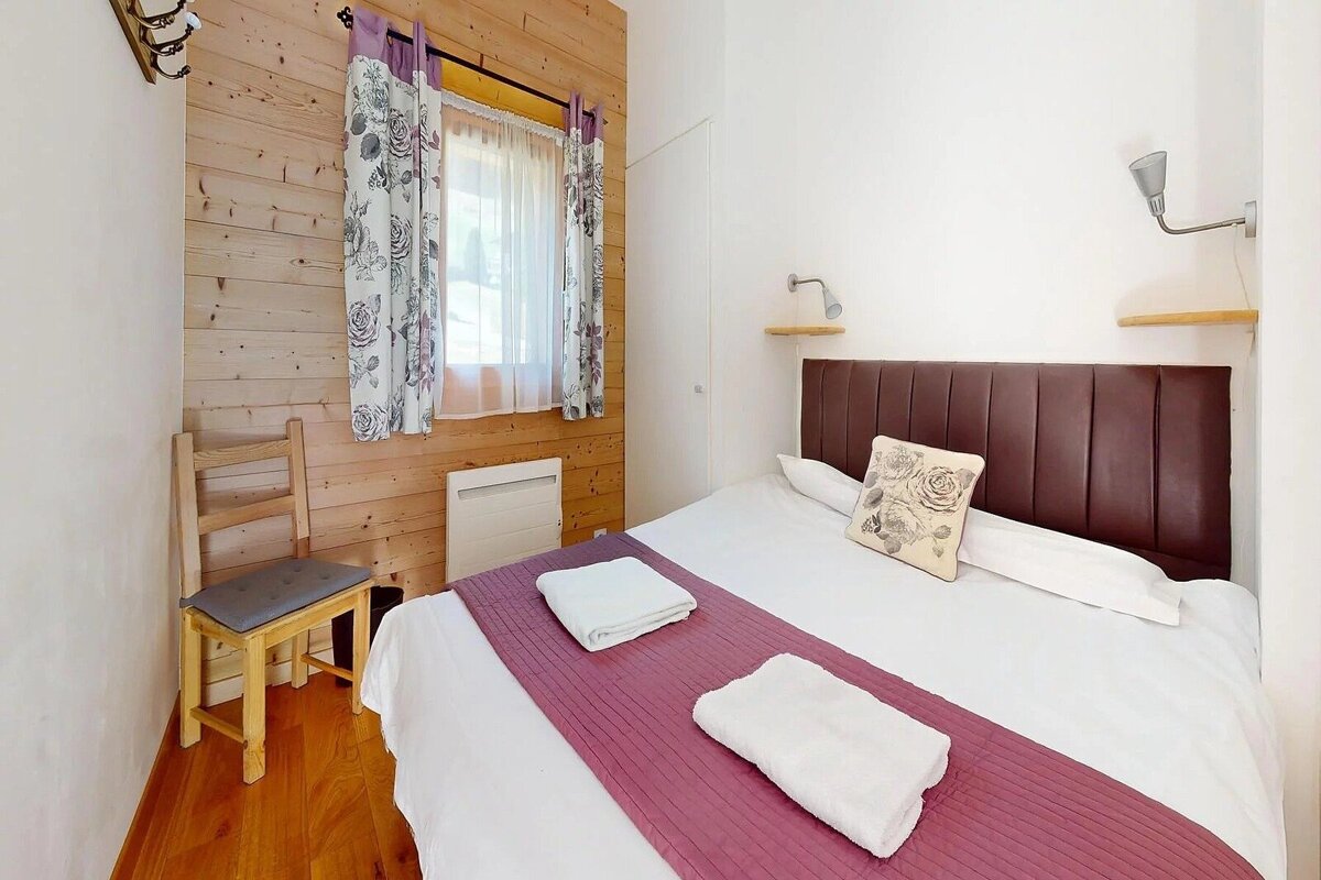 Cozy bedroom with a double bed, wood-paneled and white walls, floral curtains, a wooden chair, and reading lights above the bed.
