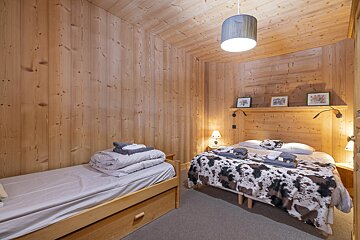 A cozy, wood-paneled bedroom featuring a main bed with a cow print blanket, a smaller trundle bed, and warm lighting, creating a rustic ambiance.