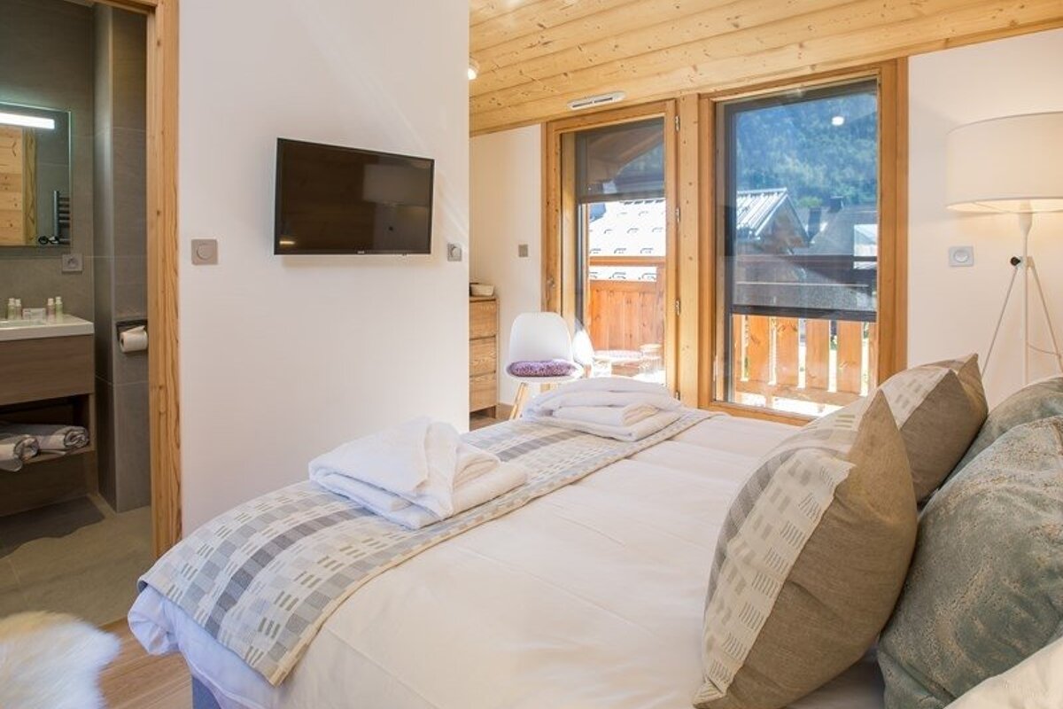 Cozy chalet bedroom featuring a neat double bed, wooden ceiling, wall-mounted TV, and large windows looking out to a balcony.