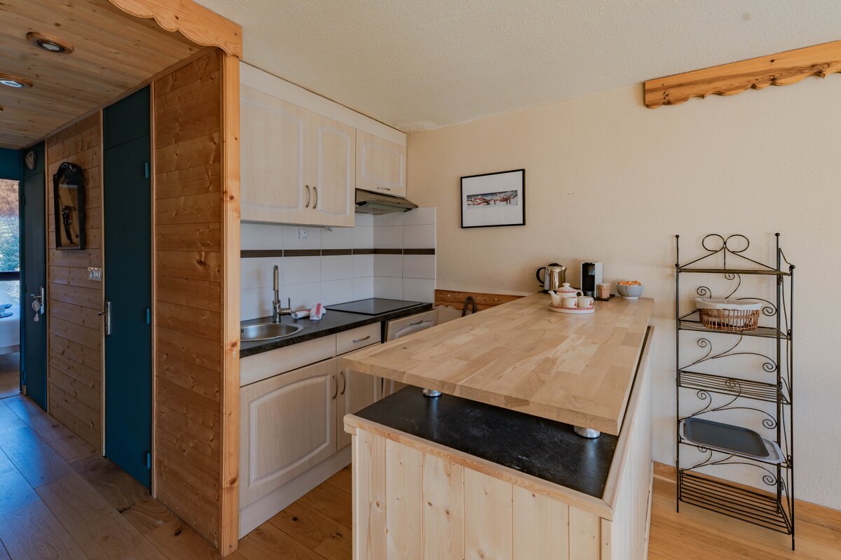 A cozy wooden kitchenette with light cabinets, a wooden breakfast bar, a metal baker's rack, and rustic wood-paneled walls.