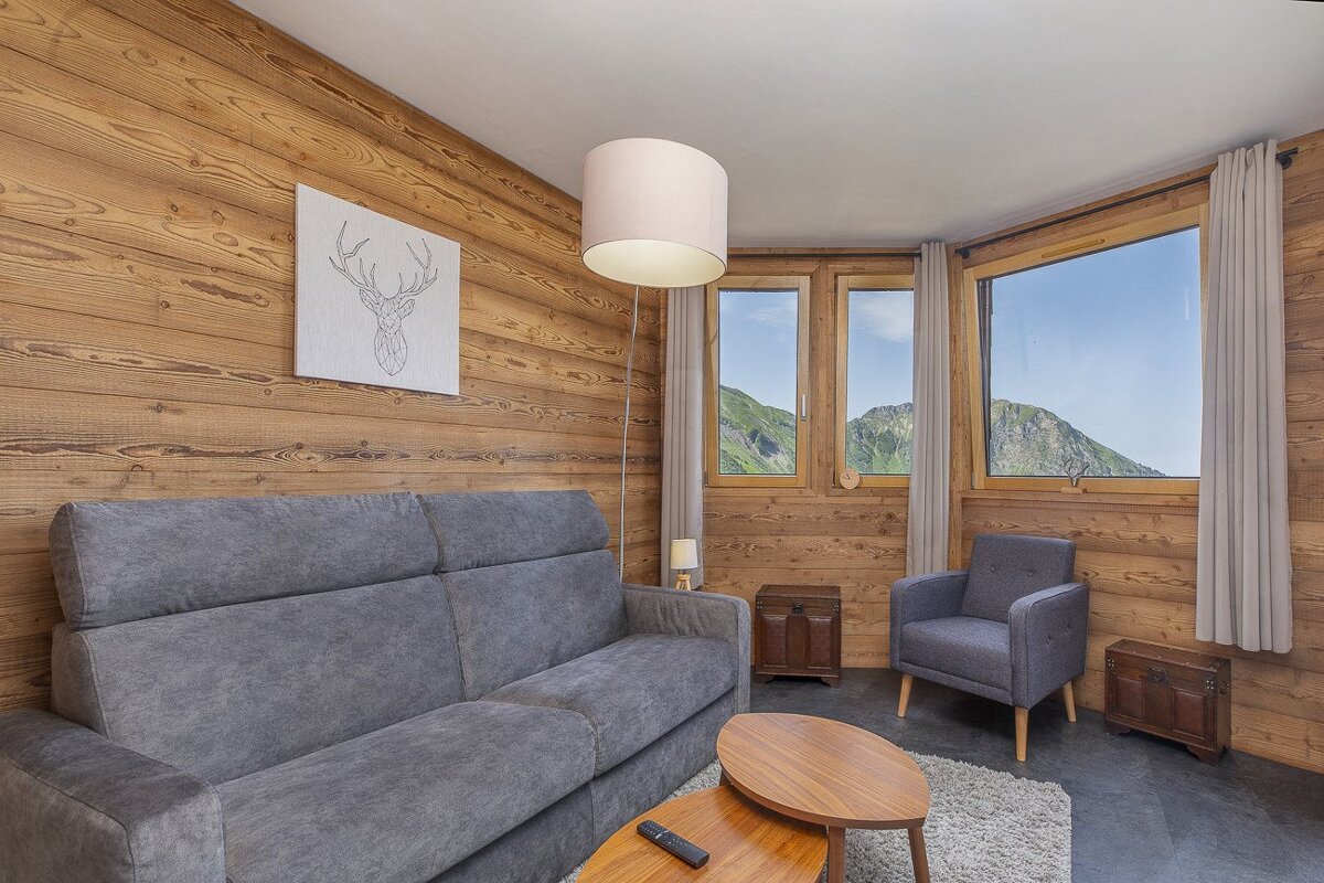 A cozy, wood-paneled living room with a grey sofa, armchair, and coffee table. Large windows offer stunning mountain views. A deer head art piece hangs on the wall.