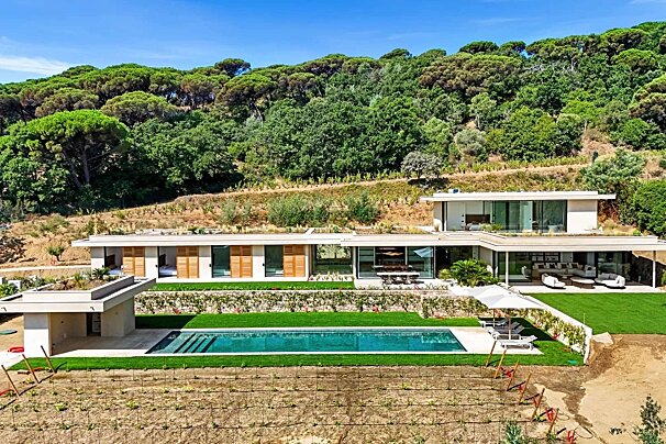 A sleek, modern villa with a long pool and green lawns, backed by a lush, forested hillside. A vineyard is visible in the foreground.