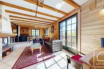 Rustic wooden chalet interior: living room with fireplace, dark sofas, exposed beams, and large windows, plus a dining area.
