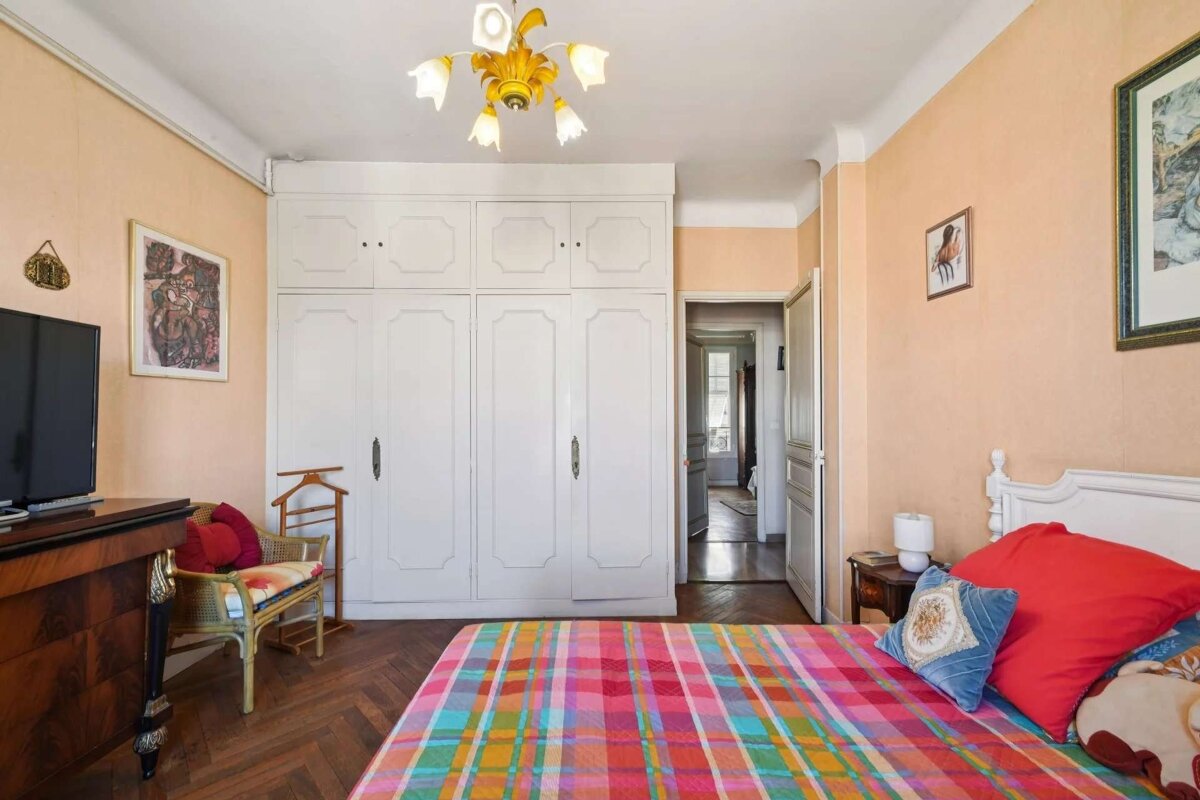 A bright bedroom with peach walls, a large white wardrobe, and a colorful plaid bedspread. It has wood floors, a white bed, and a vintage chandelier.