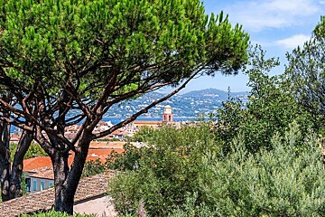 A scenic view through lush green trees of a charming coastal town with red roofs, a bell tower, and a blue sea backed by mountains.