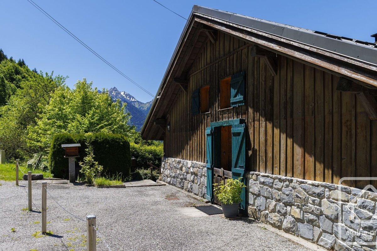 Rustic wooden house with a stone base and green shutters, set against lush trees and snow-capped mountains under a clear blue sky.