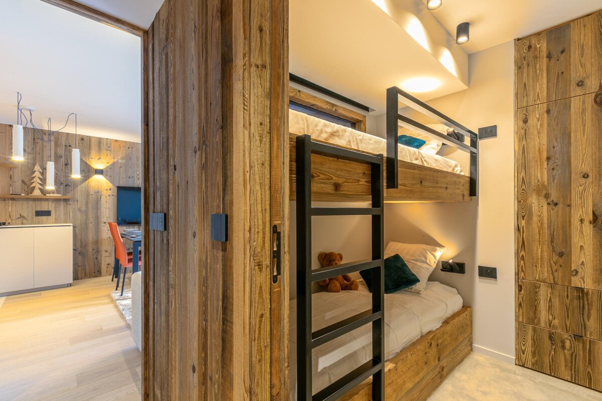 A cozy modern cabin bedroom featuring built-in wooden bunk beds, opening onto a bright dining area through a sliding wood door.