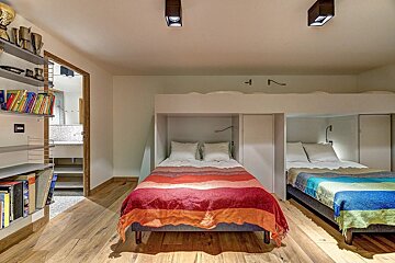 A cozy bedroom with two built-in beds featuring colorful blankets (red/orange, blue/green), two upper bunks, a wooden floor, and a bookshelf with books and trophies.