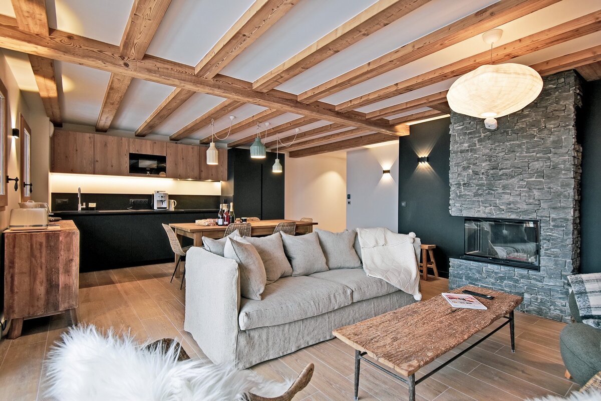 Cozy modern chalet interior featuring exposed wooden ceiling beams, a stone fireplace, a grey sofa, and a kitchen in the background.