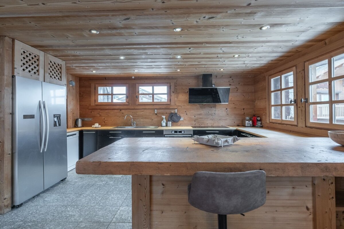 A rustic, wood-paneled kitchen with a large wooden island, a grey stool, and stainless-steel appliances.