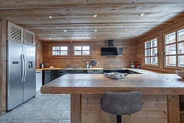 A rustic, wood-paneled kitchen with a large wooden island, a grey stool, and stainless-steel appliances.