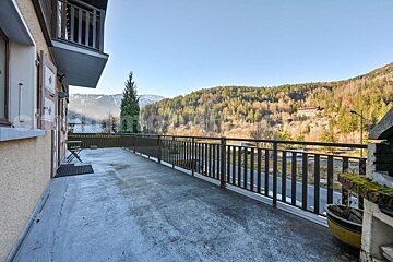 A broad concrete balcony with dark railings offers a scenic mountain vista with forests, a distant road, and snowy peaks under a clear sky.