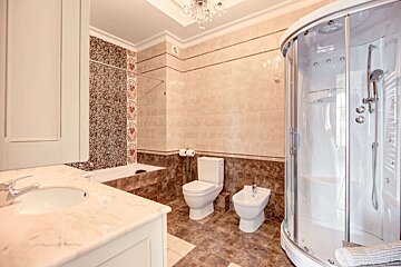 A spacious, tiled bathroom with a curved glass shower, bathtub, toilet, bidet, vanity, and a decorative chandelier on the ceiling.