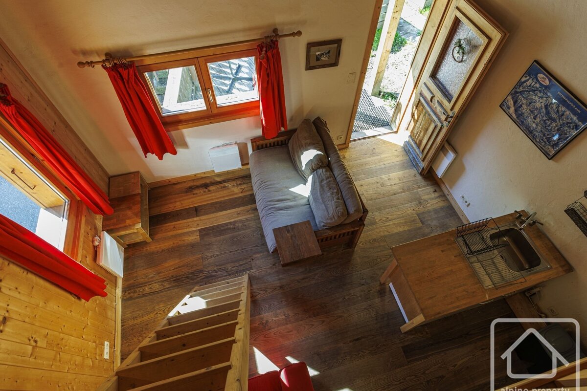 Aerial view of a rustic, cozy room with wood paneling, red curtains, a sofa, kitchenette, and stairs, bathed in sunlight.