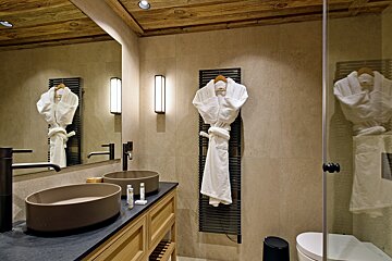 A modern bathroom with a wooden ceiling, double vanity with brown vessel sinks, black fixtures, and white robes on a towel warmer.