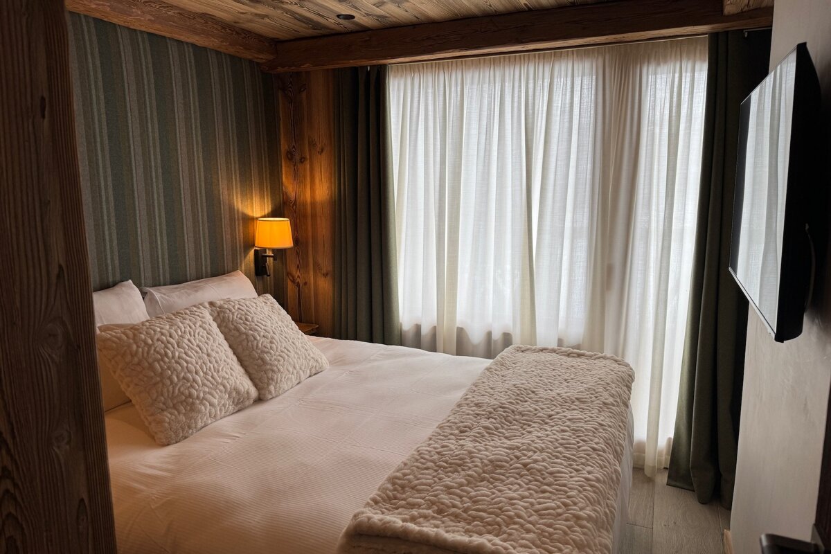 Cozy rustic bedroom with a wooden ceiling, striped wall, light bed, warm lamp, white curtains, and a wall-mounted TV.