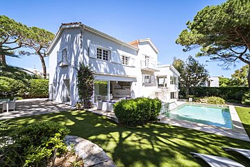 A beautiful white villa with a vibrant green lawn, a sparkling swimming pool, and tall trees under a clear blue sky.
