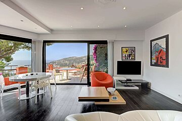 A modern, bright living room with dark wood floors and large windows opens to a balcony with stunning sea and mountain views. It features dining and lounge areas.