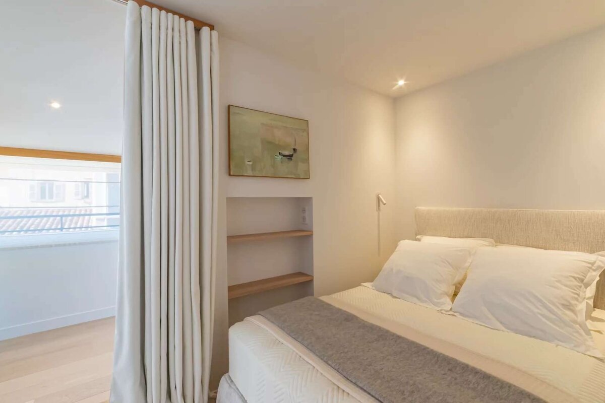 A minimalist, neutral-toned bedroom with a bed, white curtains separating a window area, built-in shelves, and abstract art. Creates a serene ambiance.
