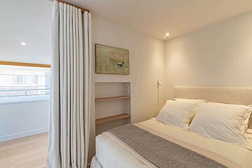 A minimalist, neutral-toned bedroom with a bed, white curtains separating a window area, built-in shelves, and abstract art. Creates a serene ambiance.