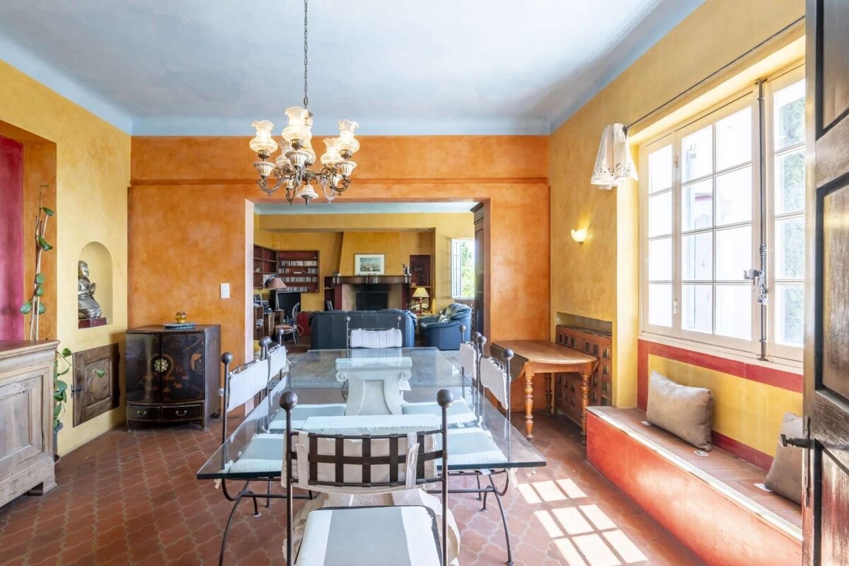 Warm, multi-colored dining room with a glass table, ornate chandelier, and terracotta floor, leading to a cozy living room. Bright window seat.