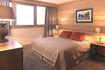 A cozy bedroom featuring wood-paneled walls, a double bed with orange pillows, a large window, plaid curtains, and warm lighting.