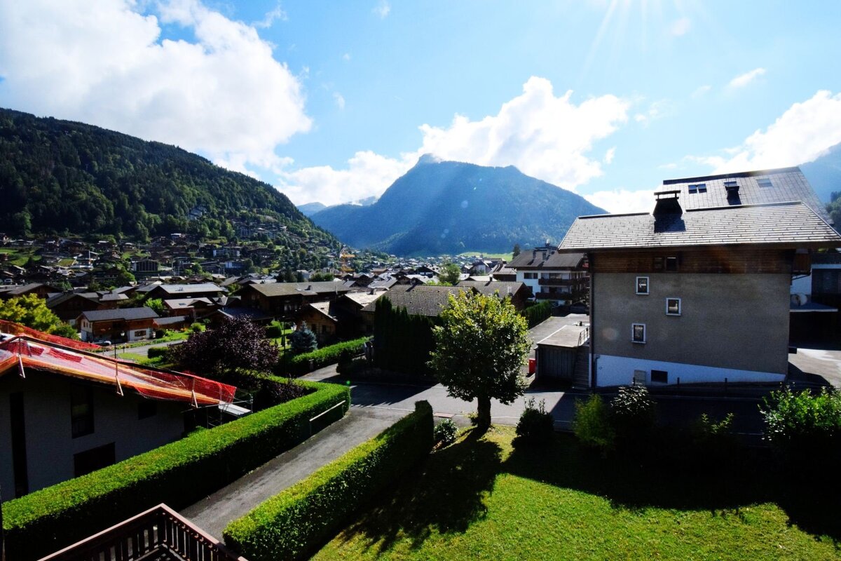 A sunny, elevated view of a European mountain village nestled in a valley with lush green hills and a bright blue sky with clouds.