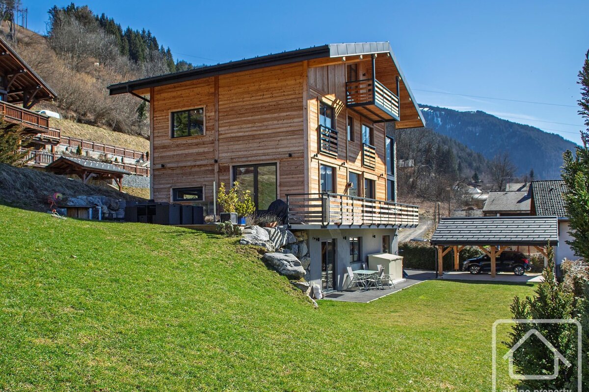 Modern wooden chalet with green lawn and mountain backdrop under a blue sky. Balconies and a carport are visible.