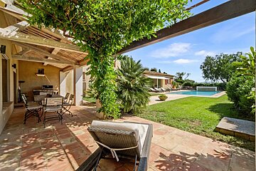 Sunny villa patio with dining furniture looking out onto a lush green lawn, palm trees, and a swimming pool.