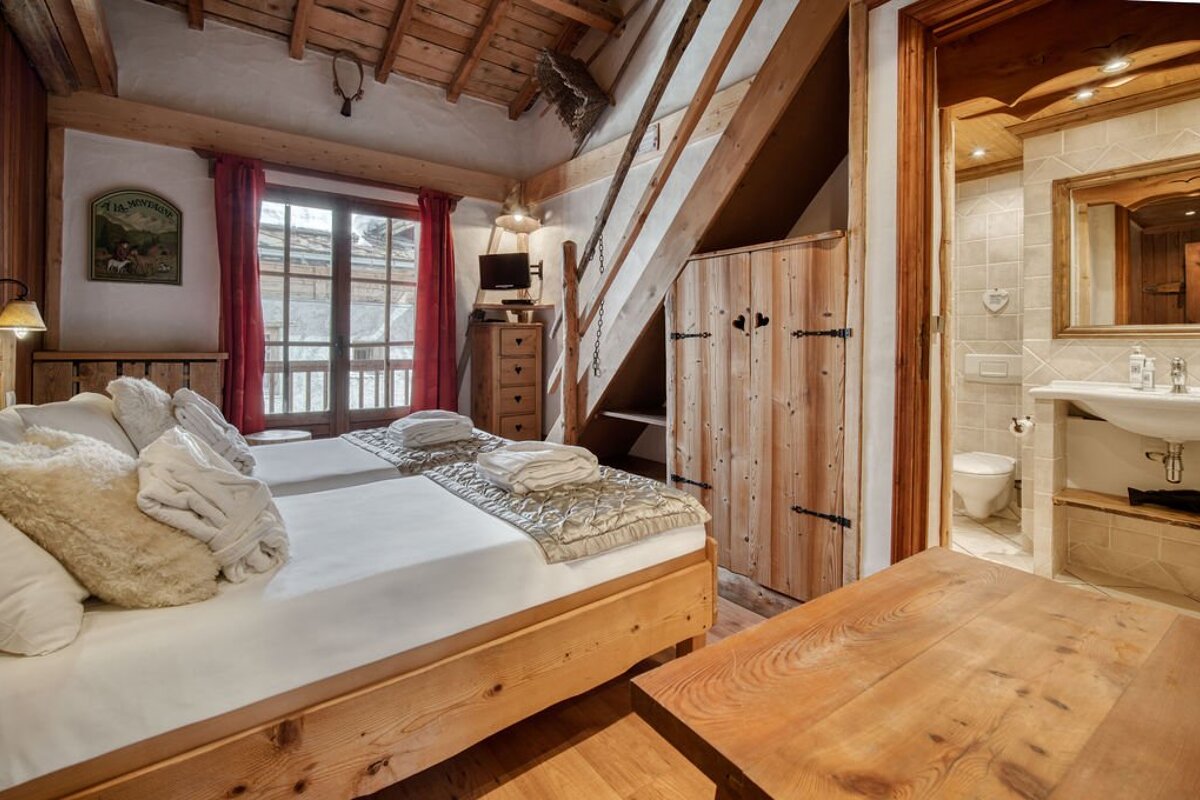 Cozy rustic chalet bedroom featuring twin beds, exposed wood beams, stairs to a loft, and a view into the adjacent bathroom.