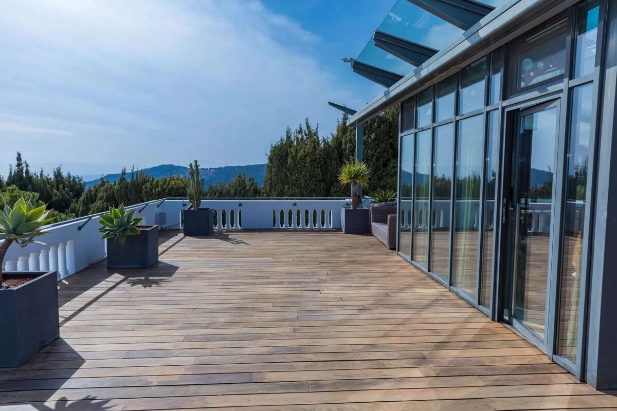 A spacious wooden deck terrace featuring a modern glass-paneled building, white balustrade, potted plants, and a scenic view of green hills.